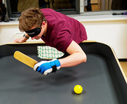 ein Spieler mit lichtundurchlässiger Brille hat einen Showdownschläger in der linken Hand und versucht den gelben Ball am Tisch zum Gegenüber zu spielen